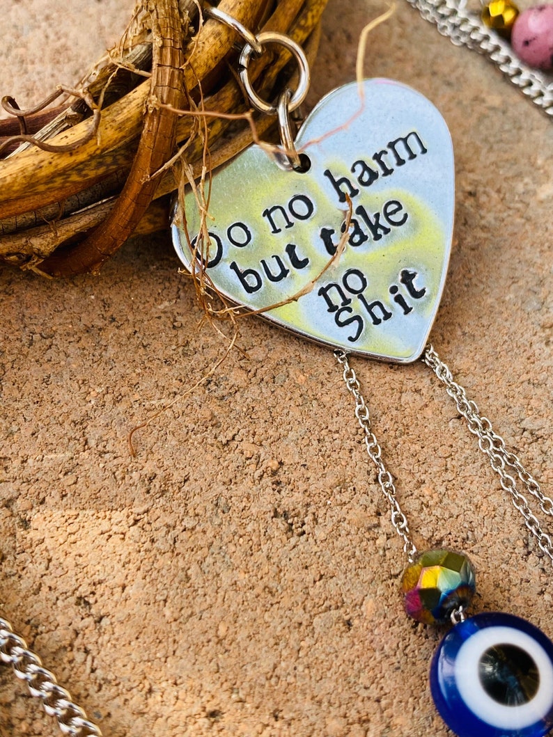 May include: A silver heart-shaped pendant with the words "do no harm but take no shit" in black lettering. The pendant hangs from a silver chain and features a blue and white evil eye charm and a faceted bead. The background is a textured, brown surface.