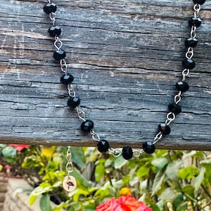 May include: A necklace featuring a series of faceted black beads linked together with silver-toned metal wire. A small, circular silver-toned charm with a detailed mushroom illustration hangs from the chain, adding a distinct focal point to the piece.