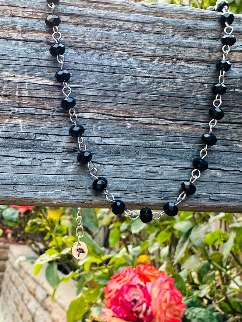 May include: A necklace featuring a series of faceted black beads linked together with silver-toned metal wire. A small, circular silver-toned charm with a detailed mushroom illustration hangs from the chain, adding a distinct focal point to the piece.