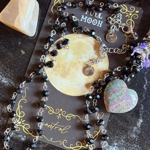 May include: A necklace featuring black faceted beads on a silver-toned chain with a heart-shaped pendant. The necklace rests on a dark card printed with a yellow moon, the text FULL MOON, and the word control in a cursive script font.