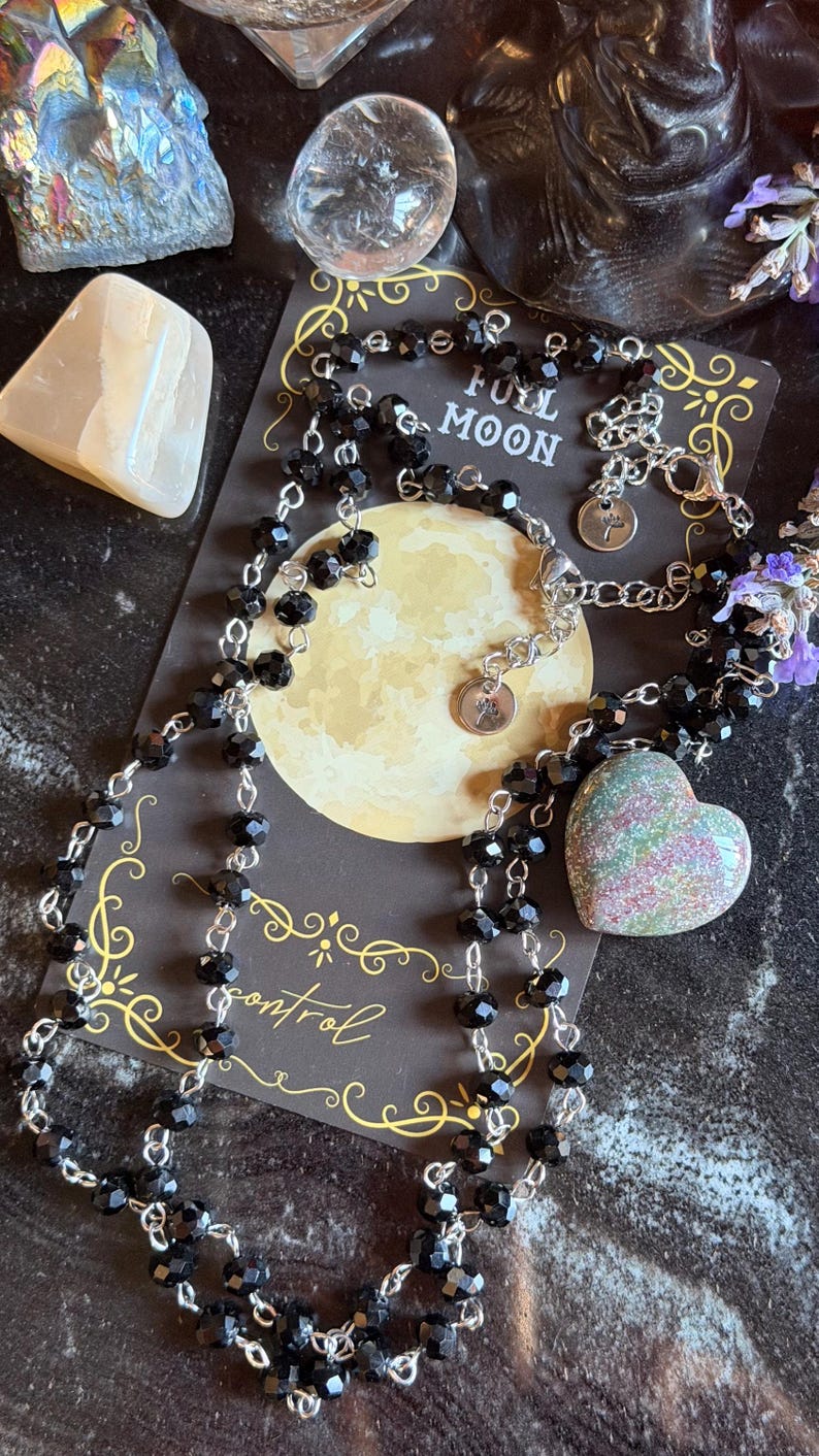 May include: A necklace featuring black faceted beads on a silver-toned chain with a heart-shaped pendant. The necklace rests on a dark card printed with a yellow moon, the text FULL MOON, and the word control in a cursive script font.