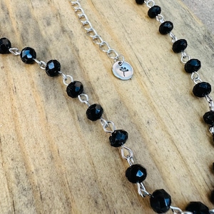 May include: A necklace featuring faceted black beads linked by silver-toned metal wire. The chain includes a silver-toned extender with a small circular charm engraved with a dandelion silhouette. The beads are uniform in size and shape.