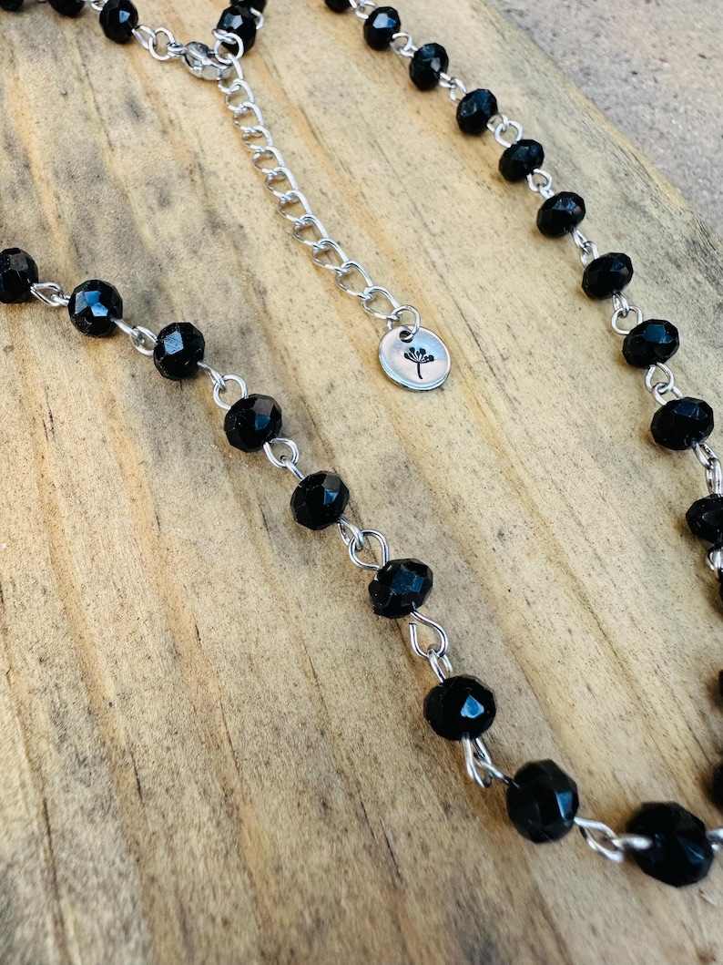 May include: A necklace featuring faceted black beads linked by silver-toned metal wire. The chain includes a silver-toned extender with a small circular charm engraved with a dandelion silhouette. The beads are uniform in size and shape.