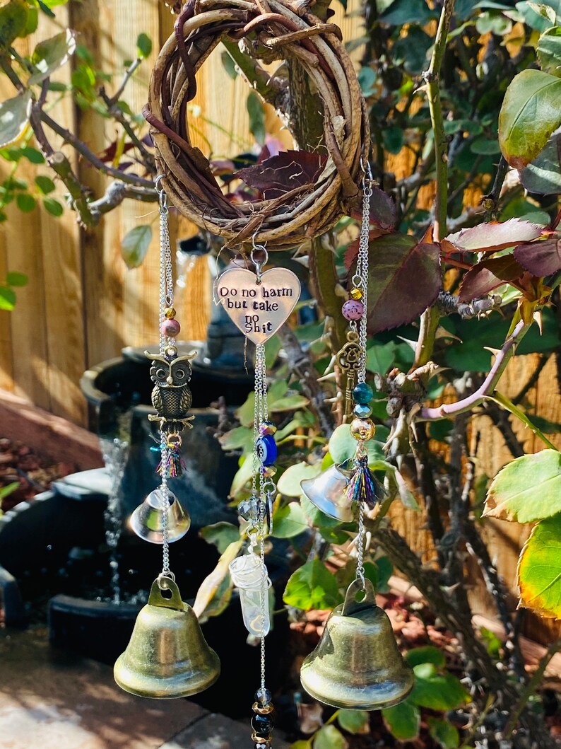 May include: A handcrafted wind chime featuring a woven grapevine ring. The chime includes a heart-shaped charm with the text "Do no harm but take no shit", along with bells, beads, and an owl charm. The wind chime is suspended from a chain.