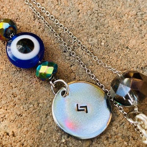 May include: Silver chain necklace with a blue evil eye bead, iridescent beads, and a circular pendant with a stamped symbol. The necklace also features a large faceted bead and a strand of white beads.