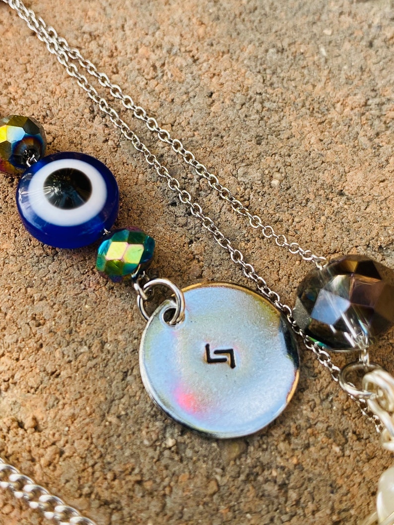 May include: Silver chain necklace with a blue evil eye bead, iridescent beads, and a circular pendant with a stamped symbol. The necklace also features a large faceted bead and a strand of white beads.