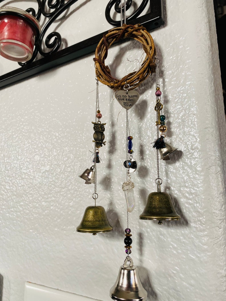 May include: A decorative wind chime featuring a woven wreath, metal bells, and various charms. The charms include an owl, beads, and a heart-shaped pendant with the text "Go no harm but take no shit". The bells are in varying sizes and finishes.