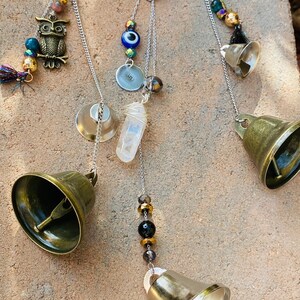 May include: A handcrafted wind chime featuring brass bells, decorative beads, and charms. The charms include an owl, a heart with the text "Do no harm but take no shit", and a clear quartz crystal. The wind chime hangs from a silver chain.