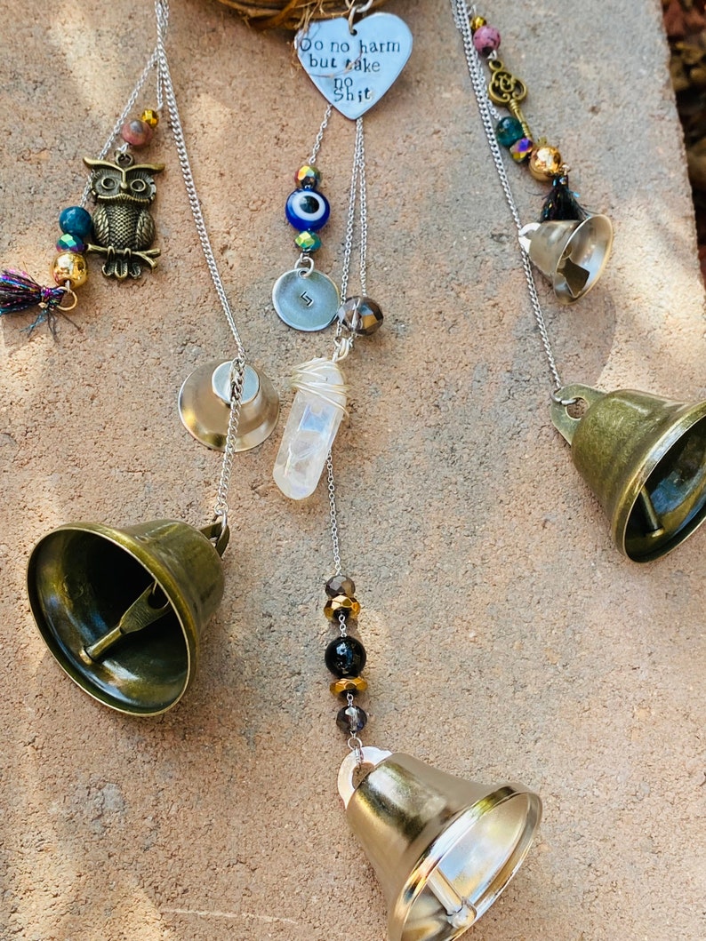 May include: A handcrafted wind chime featuring brass bells, decorative beads, and charms. The charms include an owl, a heart with the text "Do no harm but take no shit", and a clear quartz crystal. The wind chime hangs from a silver chain.