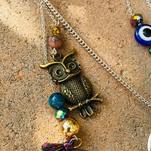 May include: A silver chain necklace with a bronze owl pendant, accented with blue, iridescent, and gold beads, and a small tassel. The owl pendant is detailed with etched feathers and eyes. The necklace is displayed on a textured, light brown surface.