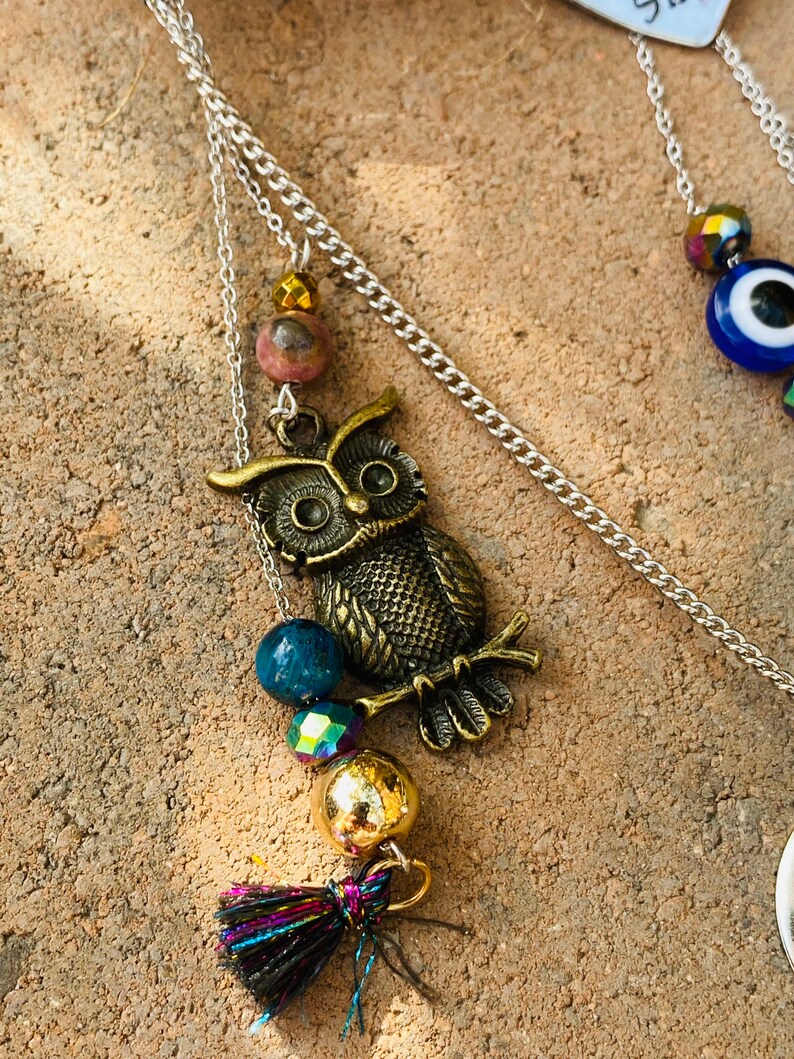 May include: A silver chain necklace with a bronze owl pendant, accented with blue, iridescent, and gold beads, and a small tassel. The owl pendant is detailed with etched feathers and eyes. The necklace is displayed on a textured, light brown surface.