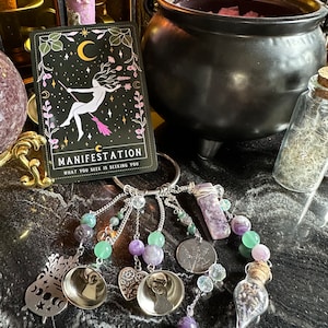 May include: A collection of mystical items, including a black cauldron, a tarot card with the word "Manifestation", and a keychain adorned with beads, charms, and a crystal. The keychain features silver-toned charms and a small glass vial.