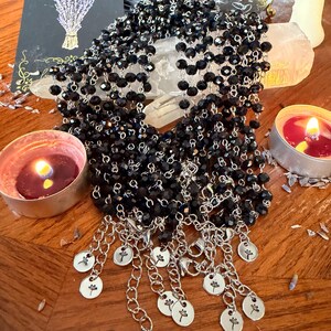 May include: A multi-strand necklace featuring faceted black beads strung on silver-toned metal chains. The necklace ends in multiple silver-toned extender chains, each finished with a small round charm engraved with a dandelion flower design.