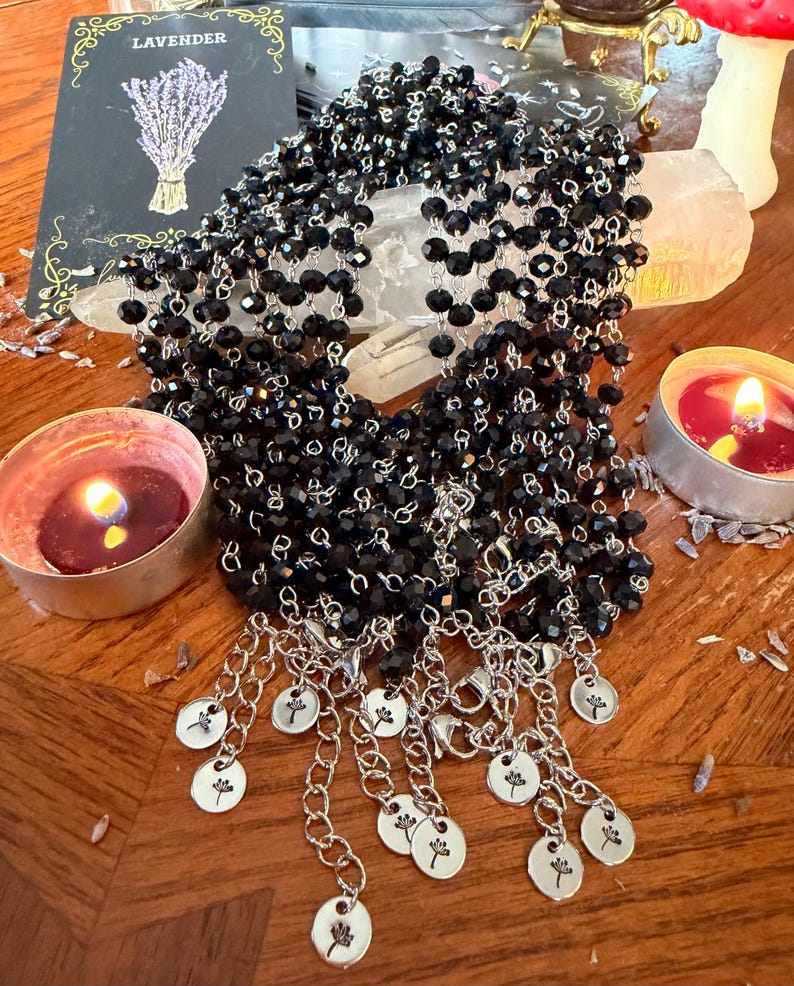 May include: A multi-strand necklace featuring faceted black beads strung on silver-toned metal chains. The necklace ends in multiple silver-toned extender chains, each finished with a small round charm engraved with a dandelion flower design.