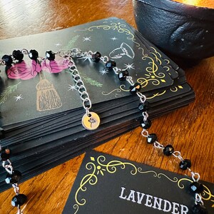 May include: A necklace featuring small, faceted black beads strung on a silver-toned metal chain. The necklace includes an adjustable extender chain ending in a small, round silver-toned charm engraved with a simple line drawing of a flower.