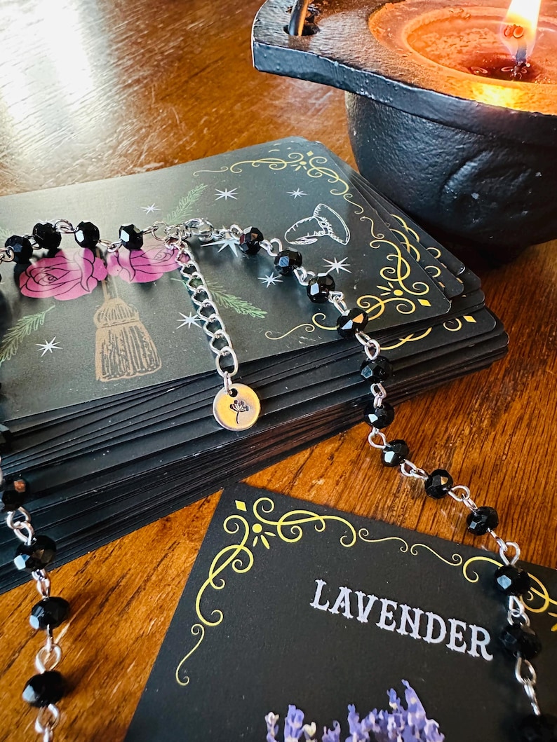 May include: A necklace featuring small, faceted black beads strung on a silver-toned metal chain. The necklace includes an adjustable extender chain ending in a small, round silver-toned charm engraved with a simple line drawing of a flower.