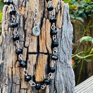 May include: A necklace featuring faceted black beads linked by silver-toned metal wire. The piece includes a silver-toned chain extender and a small, round metal charm engraved with a simple floral design, hanging from the center of the necklace.