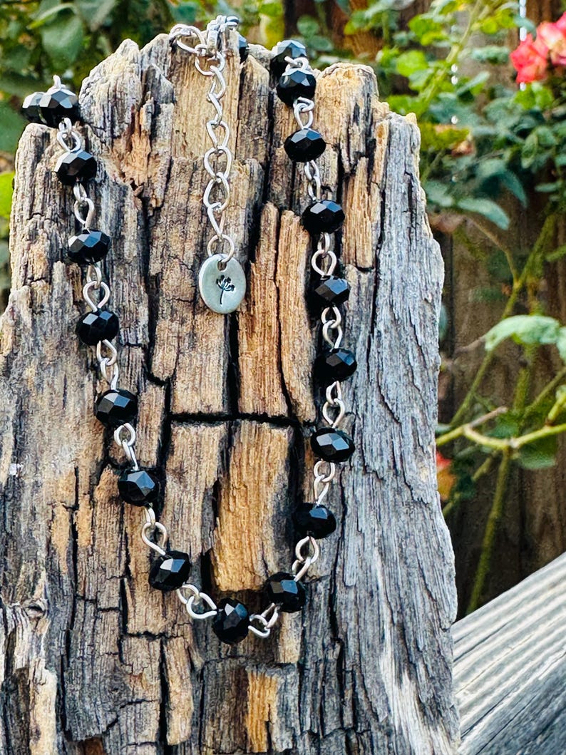 May include: A necklace featuring faceted black beads linked by silver-toned metal wire. The piece includes a silver-toned chain extender and a small, round metal charm engraved with a simple floral design, hanging from the center of the necklace.