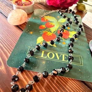 May include: A necklace featuring faceted black beads linked together with silver-toned metal wire. The beads are uniform in size and shape, creating a continuous chain design. The necklace is displayed on a green card with strawberry illustrations.