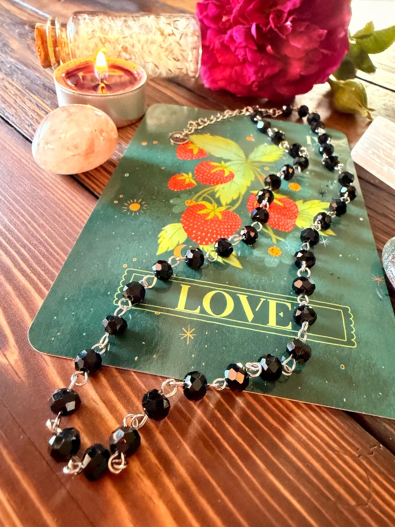 May include: A necklace featuring faceted black beads linked together with silver-toned metal wire. The beads are uniform in size and shape, creating a continuous chain design. The necklace is displayed on a green card with strawberry illustrations.