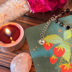 May include: A silver chain bracelet with black faceted beads and a small round charm featuring a flower engraving. The bracelet rests on a green card with a strawberry illustration, next to a lit red tea light candle and a smooth pale stone.