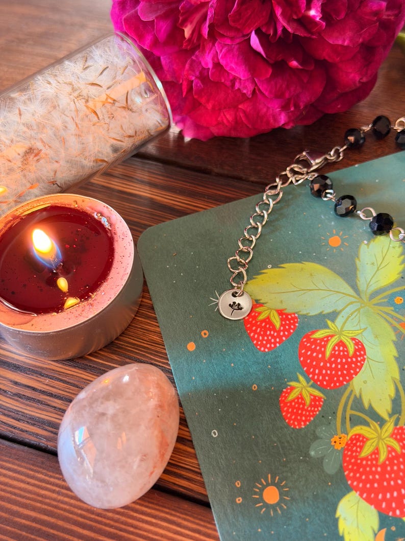 May include: A silver chain bracelet with black faceted beads and a small round charm featuring a flower engraving. The bracelet rests on a green card with a strawberry illustration, next to a lit red tea light candle and a smooth pale stone.