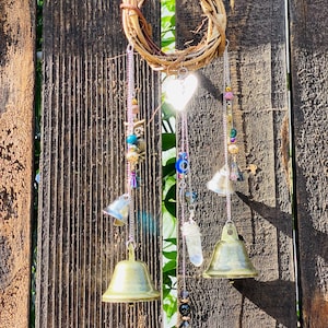 May include: A handcrafted wind chime featuring a woven grapevine wreath, hanging chains, and decorative elements. The chains support small brass bells, beads, a heart-shaped charm, and a crystal. The wind chime is suspended against a weathered wooden fence.