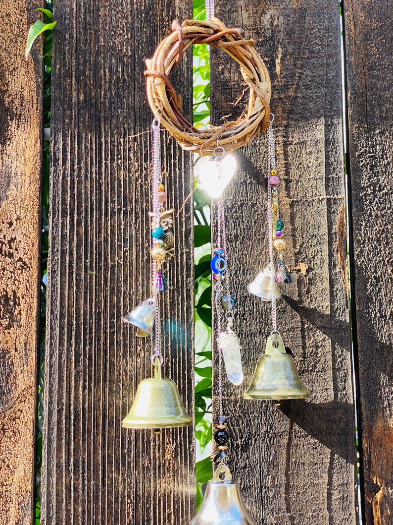 May include: A handcrafted wind chime featuring a woven grapevine wreath, hanging chains, and decorative elements. The chains support small brass bells, beads, a heart-shaped charm, and a crystal. The wind chime is suspended against a weathered wooden fence.