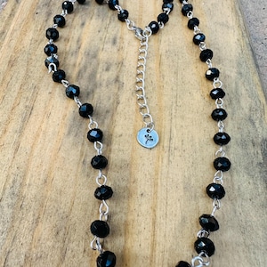 May include: A necklace featuring faceted black beads linked by silver-toned metal wire. The piece includes a silver-toned chain extender with a small circular charm at the end, which is engraved with a simple line drawing of a dandelion flower.