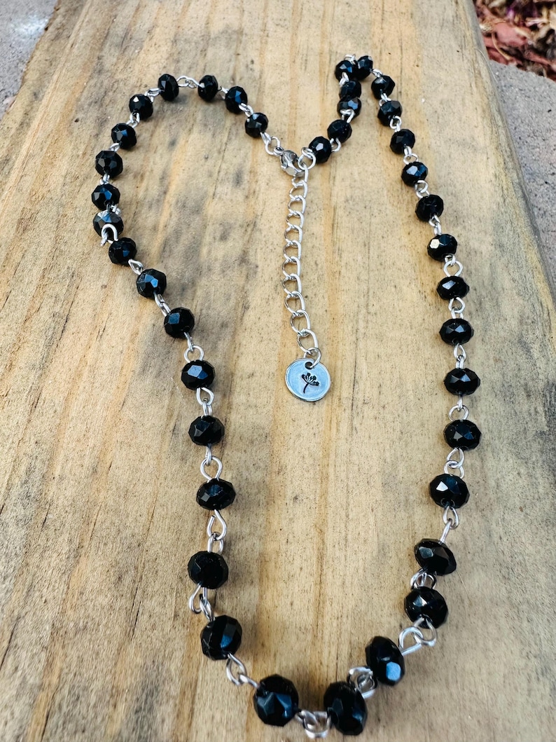 May include: A necklace featuring faceted black beads linked by silver-toned metal wire. The piece includes a silver-toned chain extender with a small circular charm at the end, which is engraved with a simple line drawing of a dandelion flower.