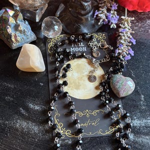 May include: A long necklace featuring faceted black beads on a silver-toned chain, displayed on a black card with a full moon graphic and the text FULL MOON and control. The necklace includes a small circular metal tag with a star symbol.