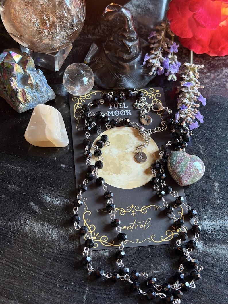 May include: A long necklace featuring faceted black beads on a silver-toned chain, displayed on a black card with a full moon graphic and the text FULL MOON and control. The necklace includes a small circular metal tag with a star symbol.