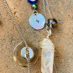 May include: A layered necklace featuring a silver chain, a clear quartz crystal wrapped in silver wire, a circular pendant with a symbol, and a blue evil eye bead. The necklace has a gold-colored circular pendant.
