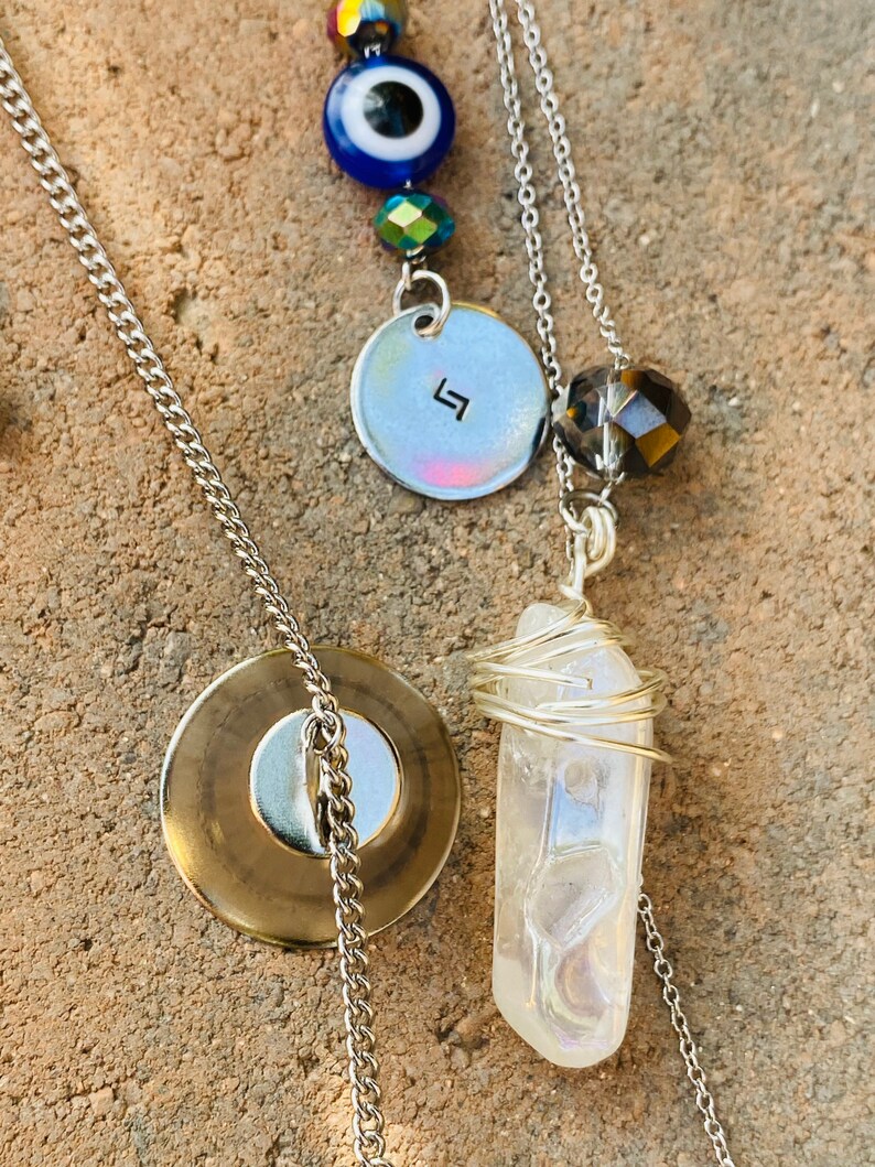 May include: A layered necklace featuring a silver chain, a clear quartz crystal wrapped in silver wire, a circular pendant with a symbol, and a blue evil eye bead. The necklace has a gold-colored circular pendant.
