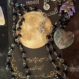 May include: A necklace featuring black faceted beads on a silver-toned chain, displayed on a black card with a full moon graphic and the text FULL MOON and control. A heart-shaped stone with green and red speckles rests beside the necklace.