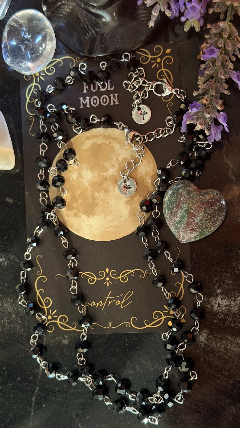 May include: A necklace featuring black faceted beads on a silver-toned chain, displayed on a black card with a full moon graphic and the text FULL MOON and control. A heart-shaped stone with green and red speckles rests beside the necklace.