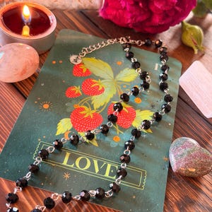 May include: A necklace featuring faceted black beads linked by silver-toned metal wire. The necklace is draped over a rectangular green card that displays a strawberry illustration and the word LOVE in gold capital letters at the bottom.