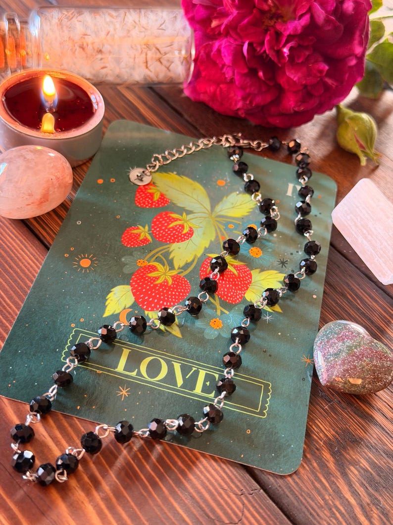 May include: A necklace featuring faceted black beads linked by silver-toned metal wire. The necklace is draped over a rectangular green card that displays a strawberry illustration and the word LOVE in gold capital letters at the bottom.