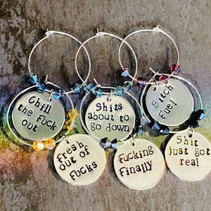 May include: Six silver wine charms, each with a different humorous phrase such as "Bitch Fuel" and "Shit just got real." The charms have beaded accents in various colours and hang from silver hoops.