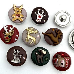 May include: A collection of circular snap buttons featuring various reindeer designs in colors including red, brown, green, and grey. Each button displays a unique reindeer motif, some with metallic finishes, small embedded crystals, or painted details.