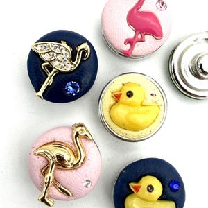 May include: A collection of five circular snap buttons featuring animal motifs. Two buttons show pink flamingos, one with gold-tone metal and one with pink enamel. Two buttons feature yellow rubber ducks on navy and cream backgrounds. One button is plain.
