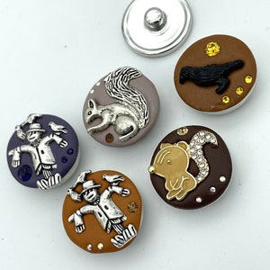 May include: A collection of decorative snap buttons with various designs. The buttons feature a silver metal base and detailed embellishments, including a squirrel, a crow, a scarecrow, and a squirrel with rhinestones. The buttons are in shades of brown, purple, and tan.