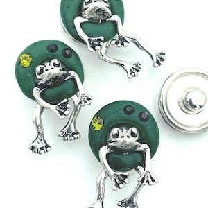 May include: A collection of frog-themed buttons. Each button features a silver-colored frog figure attached to a dark green circular base. The base is adorned with small, colorful gems. One button is detached, showing its back.
