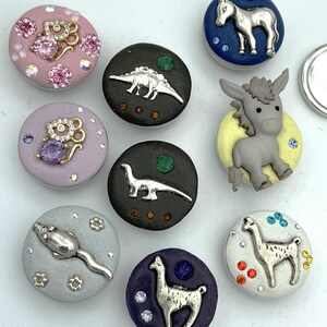 May include: A collection of decorative buttons with animal and dinosaur designs. The buttons feature silver-colored animal figures, rhinestones, and various colors, including pink, purple, black, blue, and white. The buttons are round and have a flat back.
