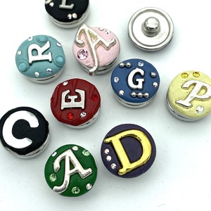 May include: A collection of circular snap buttons in various colors including black, light blue, pink, red, dark blue, yellow, green, and purple. Each button features a prominent silver or gold letter and small embedded rhinestones.