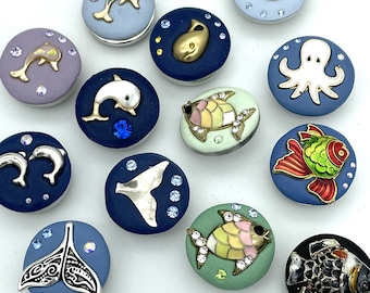 May include: A collection of 12 colorful ceramic buttons featuring various sea creatures, including fish, dolphins, whales, and an octopus. The buttons are decorated with glitter and rhinestones.