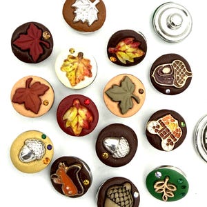 May include: A collection of circular snap buttons featuring autumn-themed designs including maple leaves, acorns, and a squirrel. Each button is decorated with small colorful rhinestones and displays various earth-toned backgrounds like brown, tan, and green.