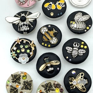 May include: A collection of decorative snap buttons with bee designs. The buttons feature various colors, including black, pink, and beige, and are adorned with rhinestones and metallic bee embellishments. Some buttons have floral and skull accents.