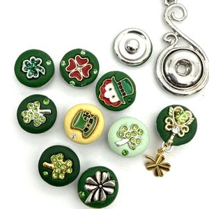 May include: A collection of circular snap buttons featuring St. Patrick's Day themes, including green shamrocks with rhinestones, a leprechaun hat, and a face. One button has a dangling gold charm. A silver-toned metal pendant is also included.