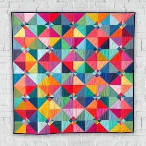 Może przedstawiać: Kolorowa kołdra z geometrycznym wzorem trójkątów w odcieniach czerwieni, pomarańczy, żółci, zieleni, błękitu, fioletu i różu. Kołdra jest obramowana ciemnoniebieskim pasem.